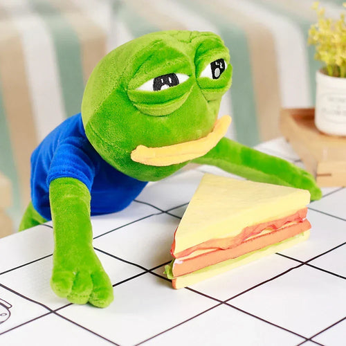 Green frog plush toy with blue shirt, lying beside a sandwich